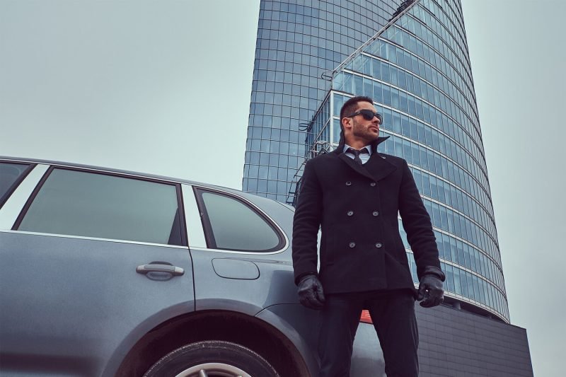 Portrait of a handsome serious male bodyguard in sunglasses standing near car outdoors.
